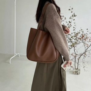 Brown large capacity leather tote bag with shoulder strap worn by a model in minimalist setting