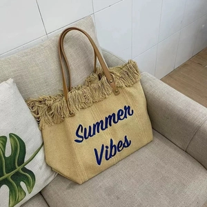 Detail of straw tote bag with blue Summer Vibes embroidery and fringe on a sofa