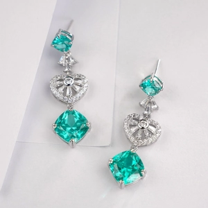 Macro detail of silver drop earrings with emerald-cut green gems and heart-shaped accents