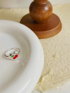 Detail of a silver open ring featuring a red cherry heart motif on white ceramic