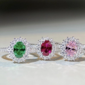 Three halo oval-cut gemstone rings in emerald green, ruby red, and pink on marble