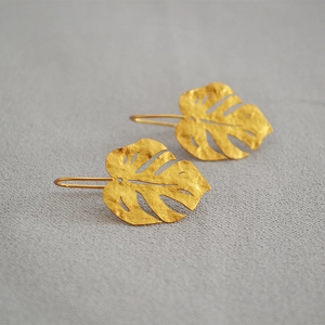 Detailed close-up of matte gold monstera leaf earrings with hammered texture on fabric