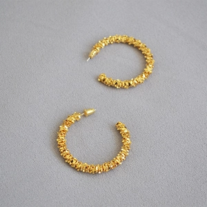 Boucles d'oreilles créoles C dorées modernes avec texture irrégulière sur tissu gris