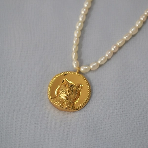 Detail of freshwater pearl necklace featuring a golden cat head coin pendant on gray fabric