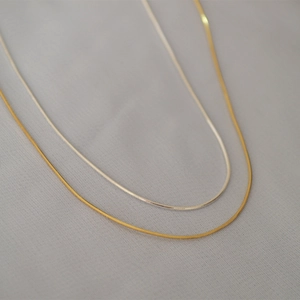 Dettaglio di collane minimaliste in oro e argento su sfondo di tessuto grigio strutturato
