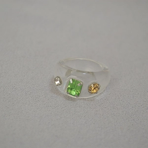 Detail of transparent resin ring featuring green rectangle and yellow round gemstones