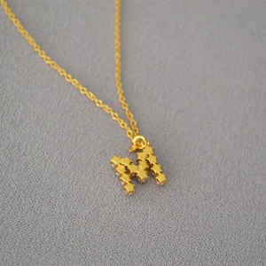 Detail des goldenen Buchstabens M mit Sternmuster-Textur und Perlen an einer feinen Kette