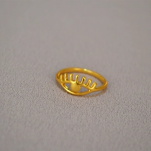 Detailed view of a gold-plated brass eye-shaped ring with eyelash cutouts on gray surface