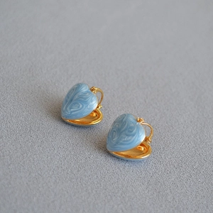 Detail of heart-shaped gold plated earrings with blue marble enamel finish on grey fabric