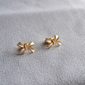 Close-up of white enamel bow earrings with gold beaded edges on a textured fabric