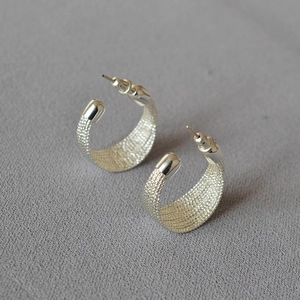 Detail of silver C-shaped stud earrings with textured surface on grey fabric background