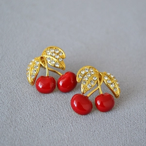 Close-up of red cherry earrings with gold leaves and sparkling rhinestones on grey fabric