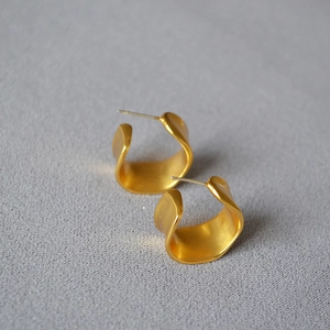 Detail of matte gold irregular C-shape wave earrings on a textured grey fabric background