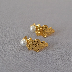 Close-up of golden leaf earrings with hammered texture and white pearls on grey fabric