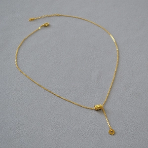 Gros plan sur le pendentif cylindre doré et le collier en Y sur un tissu gris