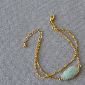 Dettaglio di bracciale dorato a doppia catena con pietra verde ovale e chiusura a moschettone