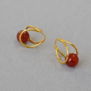 Detail of gold double-hoop earrings featuring a red agate bead on a grey fabric background