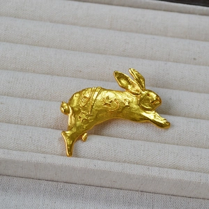 Detail of a matte gold rabbit brooch with carved star and moon motifs on a beige tray