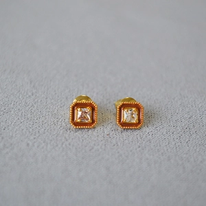 Détail de boucles d'oreilles dorées avec zircon carré et émail orange sur tissu gris