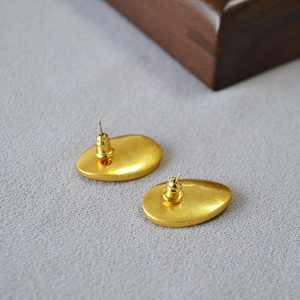 Minimalist matte gold pebble-shaped oval stud earrings on a textured grey fabric surface