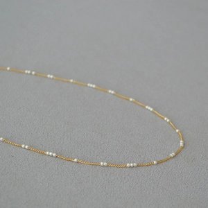 Collier minimaliste en laiton doré avec petites perles blanches sur fond texturé gris