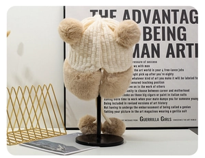 Dettaglio cappello in maglia beige con paraorecchie in peluche e quattro soffici pompon