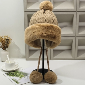 Detailed close-up of khaki cable-knit winter hat with plush pom-poms and earmuff ties