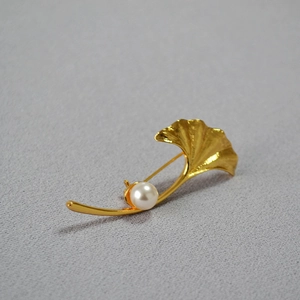 Detail of a gold ginkgo leaf brooch with a white pearl on gray textured fabric