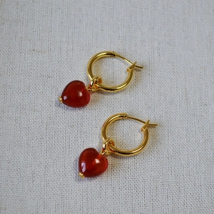 Close-up of golden hoop earrings with red heart-shaped pendants on textured fabric