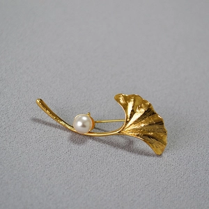 Detailed close-up of a golden ginkgo leaf brooch with a round white pearl on grey fabric