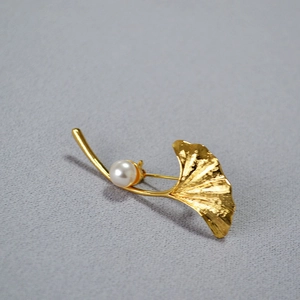 Close-up of golden ginkgo leaf brooch with white pearl on grey fabric background