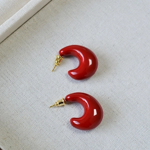 Glossy red drop C-shaped earrings with gold metal posts on a beige fabric background