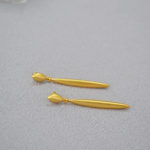 Detail of long willow-shaped matte gold earrings on a grey textured fabric background