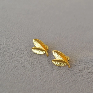 Close-up of golden leaf stud earrings with detailed matte metal texture on grey fabric