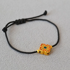 Detail of adjustable black woven bracelet featuring a gold flower charm with red enamel