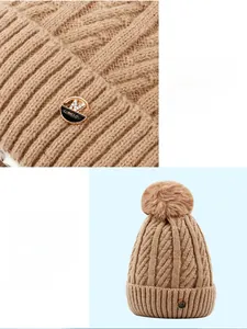 Detail of cable knit pattern and metal logo on dark camel winter beanie with pompom