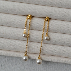Detail of long gold earrings with dual metal chains and round white pearls on a tray