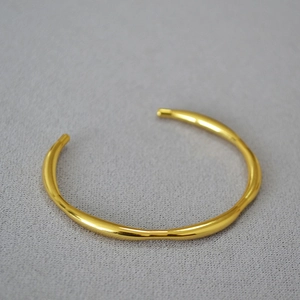 Detailed top view of gold-plated open bangle with irregular wavy lines on grey fabric