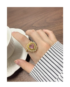 Détail d'une bague coeur dorée avec zircon rose et perles sur une main tenant une tasse