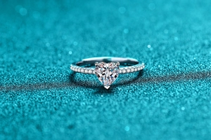 Macro detail of a silver heart-shaped moissanite ring with pavé diamonds on a blue background