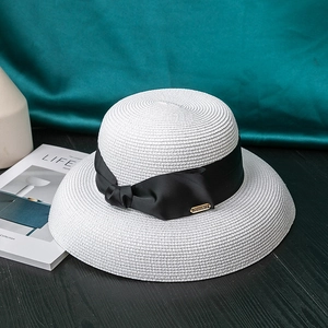 Detail of white woven straw hat featuring a black satin ribbon bow and gold metal tag