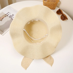 Detail of beige straw hat featuring wavy wide brim and elegant bow on white table