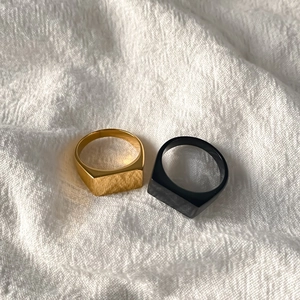 Close-up of minimalist square signet rings in matte gold and black finish on white fabric