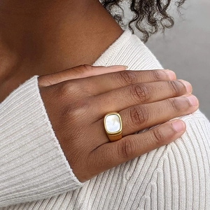 Gros plan d'une bague carrée en acier doré avec pierre blanche sur une main
