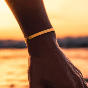 Minimalist gold stainless steel open bangle worn on wrist with water droplets on beach