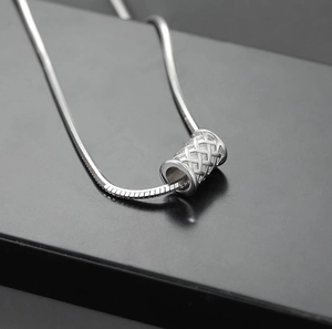 Macro shot of silver snake chain necklace with hollow woven cylinder pendant on black stand