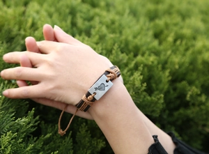 Macro detail of brown braided leather bracelet featuring an engraved heart and arrow charm