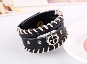 Détail macro du bracelet en cuir noir avec badge croix argenté et couture blanche à la main