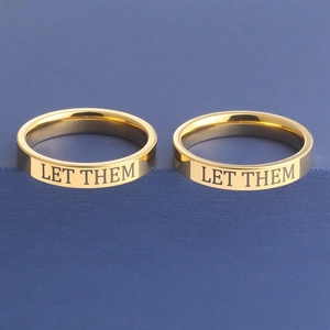 Detail of two gold titanium steel rings with Let Them engraving on blue texture