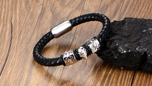 Detail of woven black leather bracelet featuring a silver skull and cross sliders on wood